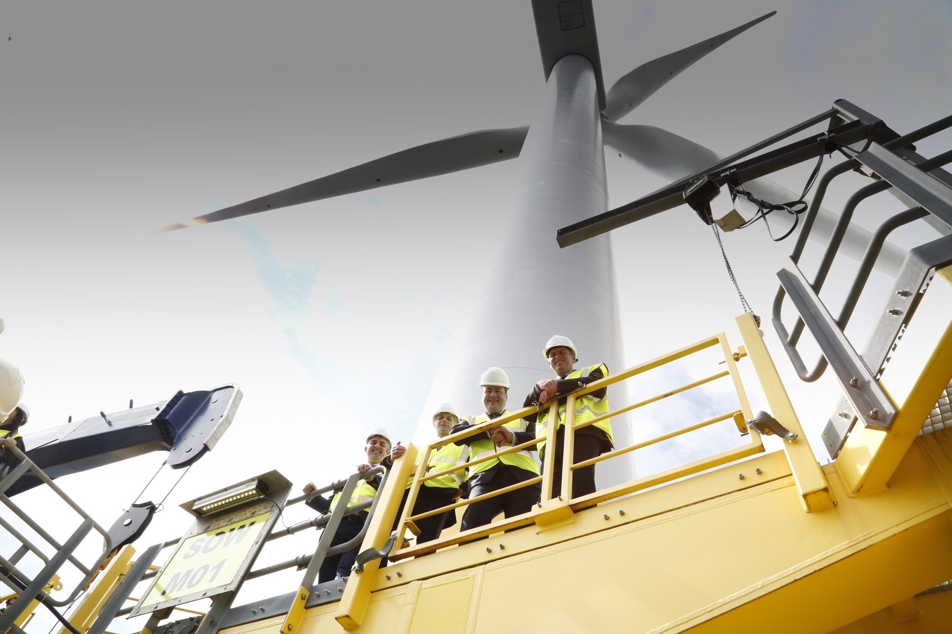Workmen stand on the base platform of a wind turbine and look down, the wind turbine looms in the background.