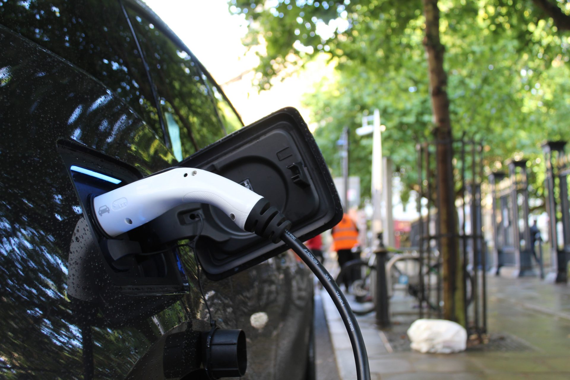 A charging cable is plugged into an electric car parked at the side of the road. Green trees in the background.