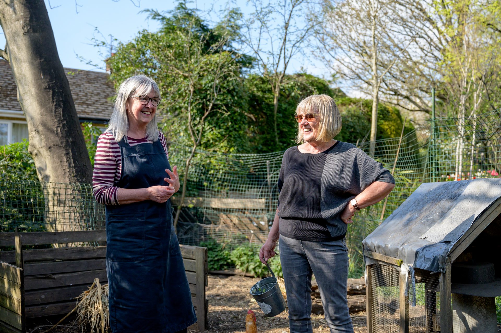 Two elderly women are standing in a garden in the sun, laughing and chatting.