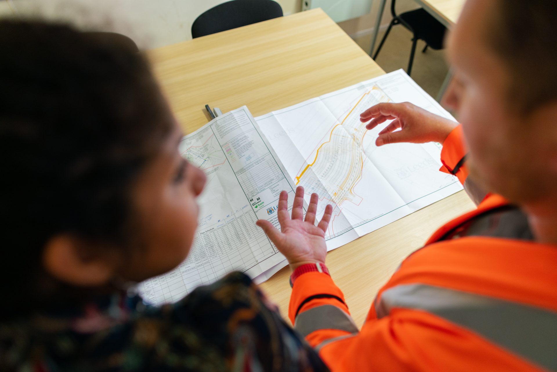 Two people are standing in front of a planning map on the table. One of them is wearing an orange high-visibility vest.